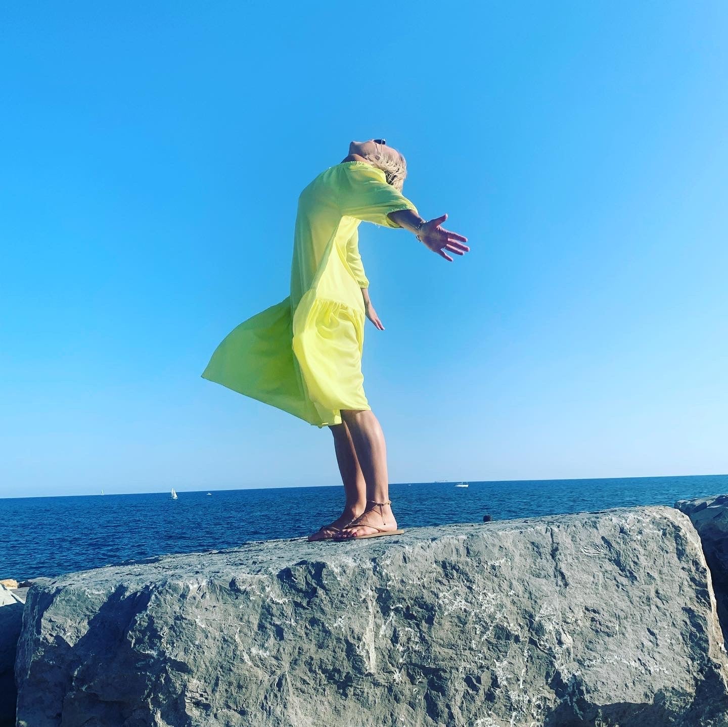 Woman standing on rock by sea in yellow dress reaching for the sky