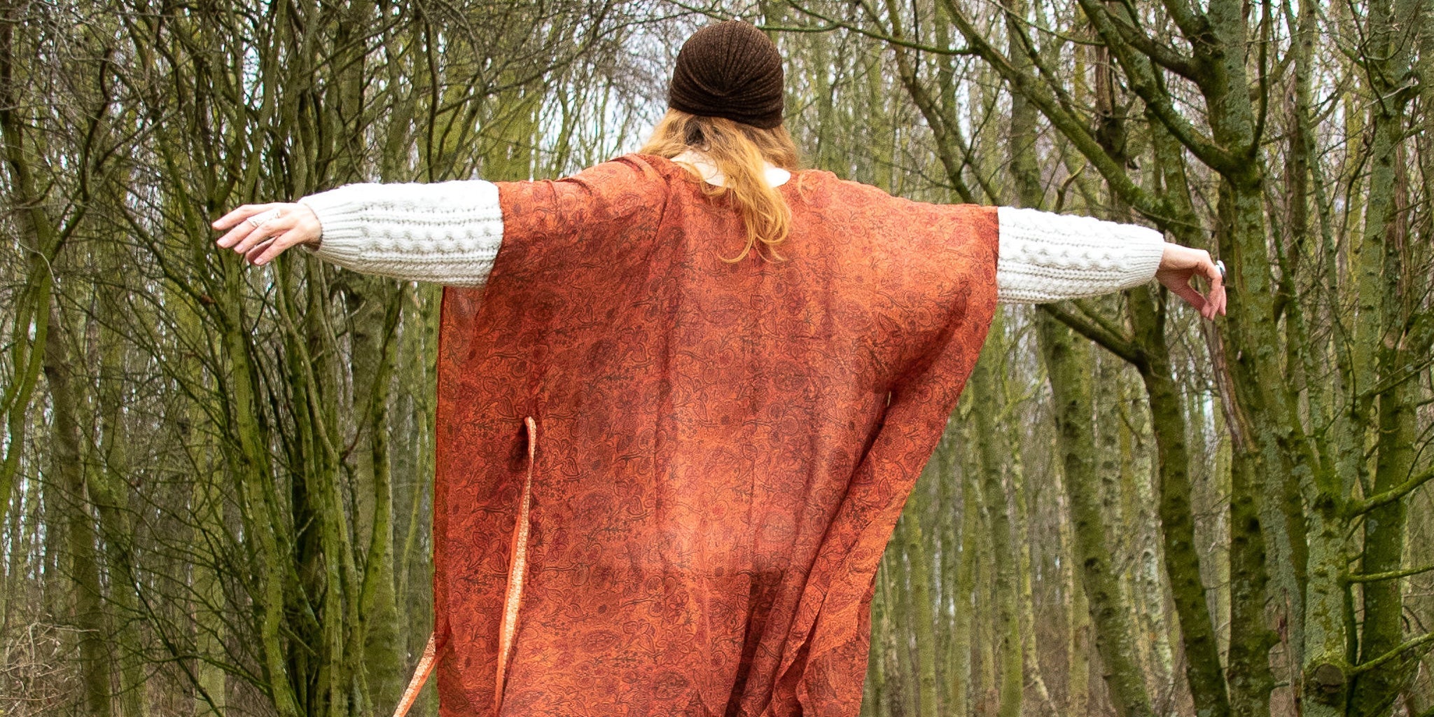 Woman in woods wearing neem kimono style wrap dress in ochre.  Her back to you with arms outstretched