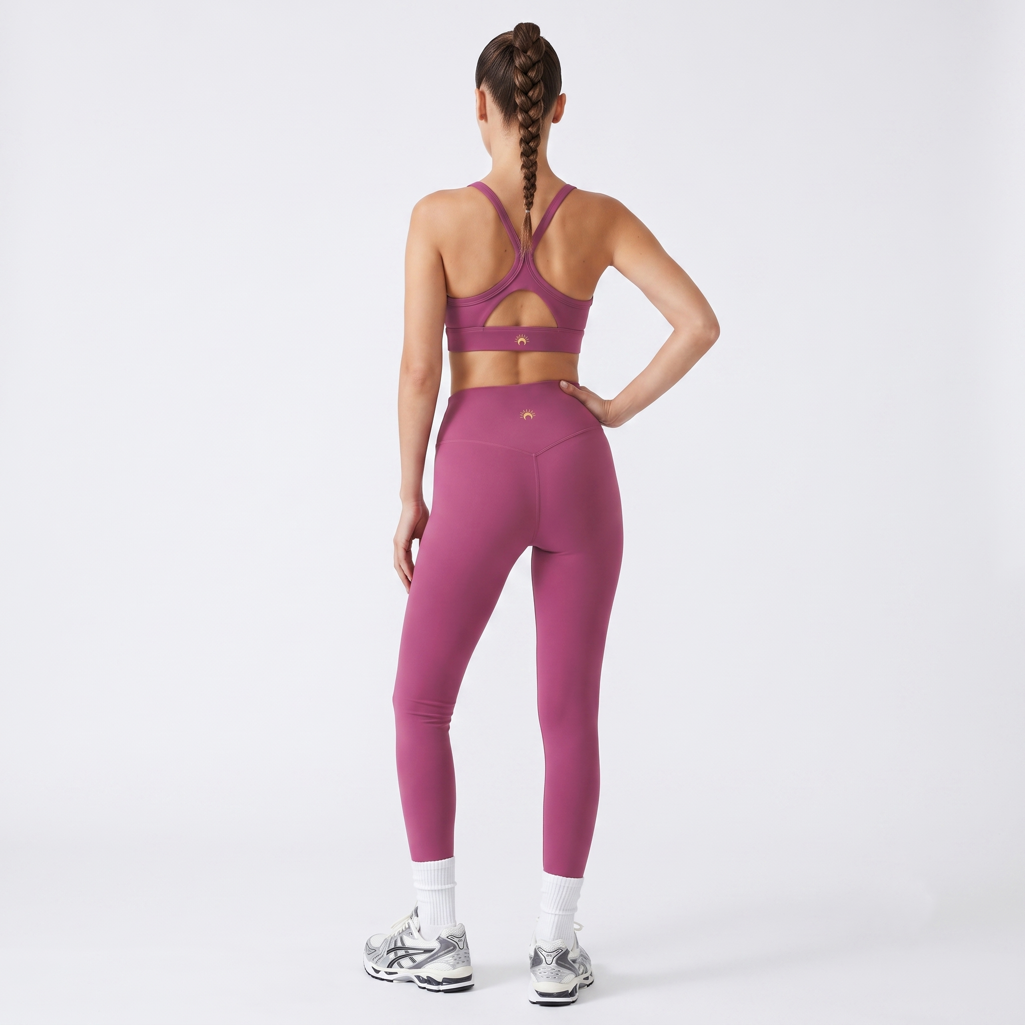 A woman shown from the back wearing a matching mauve-pink sports bra and high-waisted leggings set, paired with white socks and silver sneakers, against a light gray studio background.