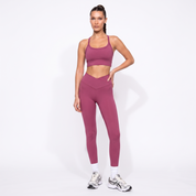 A woman modeling a matching dusty rose activewear set consisting of a sports bra and high-waisted leggings, paired with white socks and athletic sneakers, against a plain white studio background.