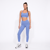A woman modeling a matching light blue activewear set consisting of a sports bra and high-waisted leggings, paired with white socks and silver sneakers, against a plain white studio background.