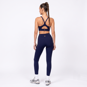 A woman stands with her back to the camera wearing a navy blue sports bra with an X-back cutout and matching high-waisted navy blue leggings, paired with white socks and silver sneakers, against a plain white studio background.