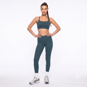 A woman modeling a matching forest green activewear set consisting of a sports bra and high-waisted leggings, paired with white socks and athletic sneakers, against a plain white studio background.