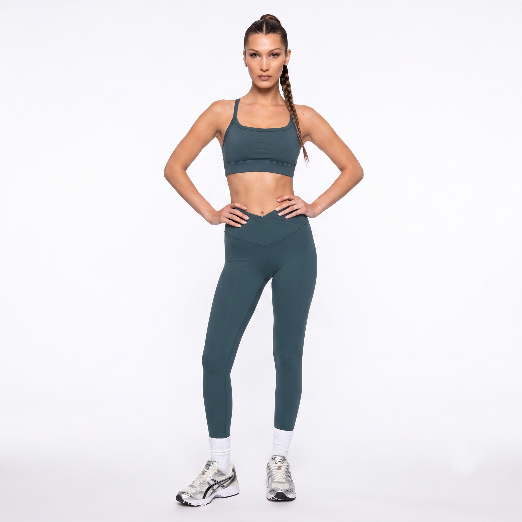 A woman modeling a matching forest green activewear set consisting of a sports bra and high-waisted leggings, paired with white socks and athletic sneakers, against a plain white studio background.